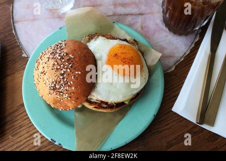 Hamburger caldo con il lato soleggiato in alto uovo su un brioche con semi di papavero e tutto il condimento su un piatto di colore teal. Foto Stock