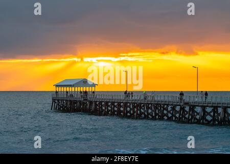 Adelaide, Australia - 11 aprile 2021: Persone che camminano lungo l'Henley Beach Jetty mentre guardano il tramonto drammatico e tempestoso Foto Stock