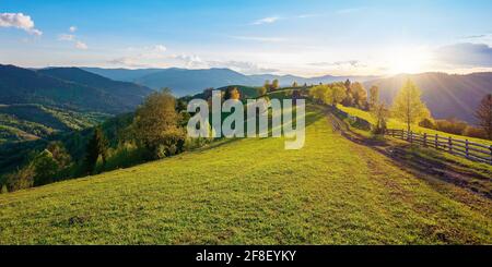 alberi dietro la recinzione sul prato erboso. primavera paesaggio rurale in luce sera. lontano crinale di montagna sotto un cielo luminoso con nubi soffici Foto Stock