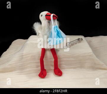 Immagine concettuale di come covid-19 influenza la gravidanza e il feto. Cicogna con fascio di maschera facciale e siringa con ago Foto Stock