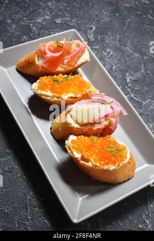 Assortimento di bruschette con baguette, formaggio cremoso, caviale rosso, salmone, aringhe e cipolla serviti su fondo di pietra nera. Set di antipasti con Foto Stock