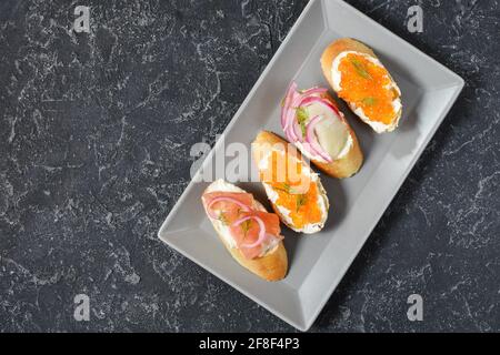 Assortimento di bruschette con baguette, formaggio cremoso, caviale rosso, salmone, aringhe e cipolla serviti su fondo di pietra nera. Set di antipasti con Foto Stock