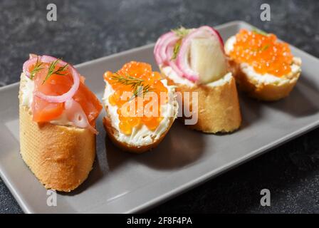 Assortimento di bruschette con baguette, formaggio cremoso, caviale rosso, salmone, aringhe e cipolla serviti su fondo di pietra nera. Set di antipasti con Foto Stock