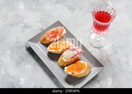 Assortimento di bruschette con baguette, formaggio cremoso, caviale rosso, salmone, aringhe e cipolla serviti su fondo di cemento. Set di antipasti con var Foto Stock