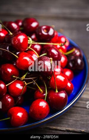 Ciliegie mature e succose in piatto blu su fondo rustico scuro. Messa a fuoco selettiva. Profondità di campo poco profonda. Foto Stock