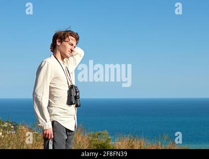 Ritratto di un giovane stanco con binocolo in piedi di fronte a uno scenario di mare che guarda lontano Foto Stock