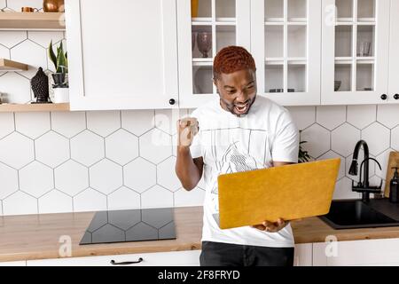 Un freelance o uno studente che si è accanito con grande gioia sta usando un computer portatile, si sente euforico, ha ottenuto una promozione di lavoro o ha superato esami importanti. Eccitato uomo d'affari afroamericano Foto Stock