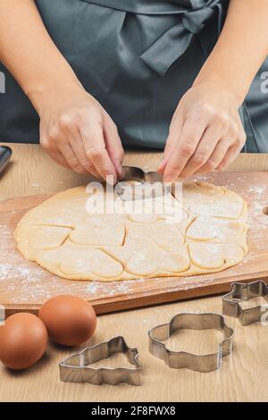 Le mani delle donne tengono una taglierina metallica a forma di cuore e tagliano i biscotti dall'impasto. Primo piano, vista laterale. Foto Stock