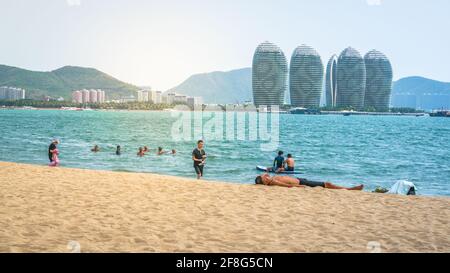 Sanya Cina, 24 marzo 2021 : persone che si adagiano sulla spiaggia di Sanya e gli edifici dell'isola di Phoenix vista nella baia con la luce drammatica in Sanya Hainan CH Foto Stock