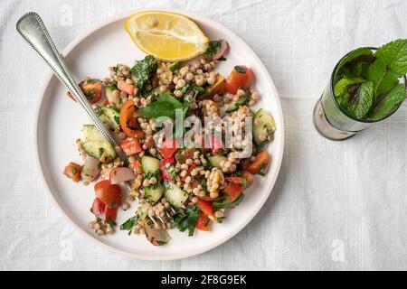 Flatlay di un piatto unico di un sano grano integrale vegano insalata couscous perlato con limone e verdure fresche su un piatto bianco servito con tè alla menta Foto Stock