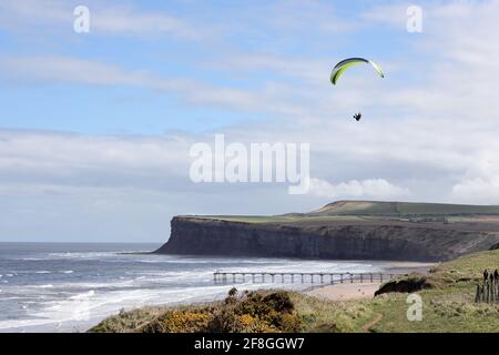 Parapendio volando lungo la costa con il molo di Saltburn e le scogliere di Huntcliff sottostanti, Saltburn-by-the-Sea con il North Yorkshire, Regno Unito Foto Stock