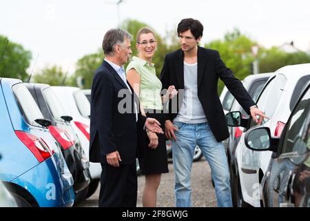 Coppia rivenditore di auto e coppia giovane in piedi sul posto di parcheggio in concessionaria nella parte anteriore delle vetture Foto Stock