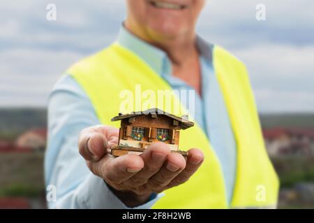 Messa a fuoco selettiva primo piano di minuscoli case di legno tenuto da vecchio costruttore maschio che indossa gilet fluorescente da lavoro con sfondo casa Foto Stock