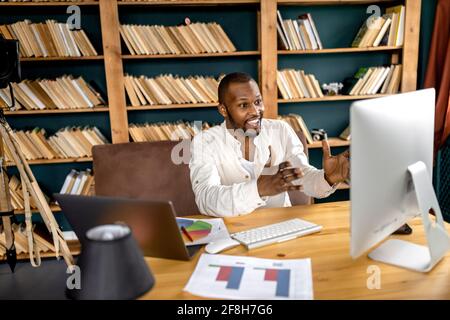 Gioioso freelance maschile sta usando il computer, discutendo idee con i colleghi in video riunione, sorridendo. Uomo d'affari riuscito sta avendo un in linea Foto Stock