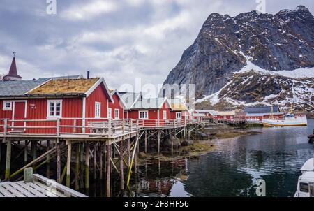 Tipiche case rosse di Reine Fishing Village, Isole Lofoten, Norvegia. Nessuna gente. Porto con barche e montagna in neve. Foto Stock