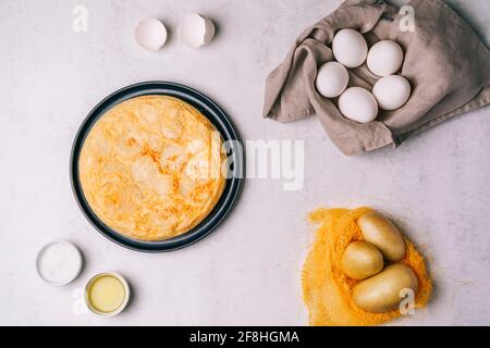 Vista aerea di una frittata spagnola su un piatto nero su un tavolo di legno con gli ingredienti sul lato, uova, patate, olio d'oliva e sale Foto Stock