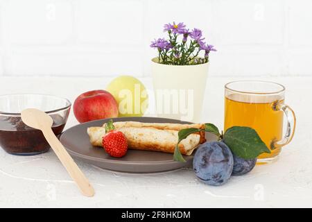 Frittelle fatte in casa farcite di formaggio casereccio, una fragola su un piatto, una ciotola di vetro con marmellata, una tazza di tè, prugne, mele e fiori. Foto Stock