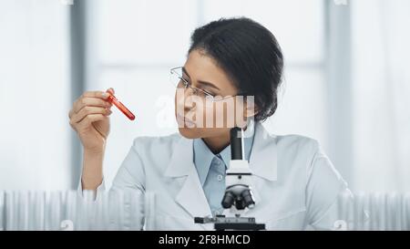 scienziato afroamericano in bicchieri che tengono una provetta con sangue campione vicino al microscopio in laboratorio Foto Stock