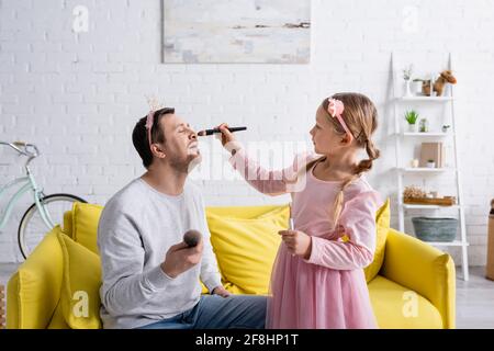 figlia che applica il trucco sulla faccia del padre che siede sul divano nella corona giocattolo Foto Stock