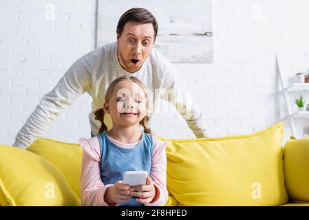 uomo sorpreso che guarda lo smartphone nelle mani di figlia seduta sul divano Foto Stock