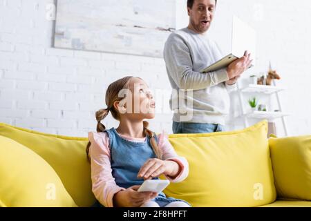 uomo sorpreso con un computer portatile che guarda la figlia che copre lo smartphone con mano Foto Stock