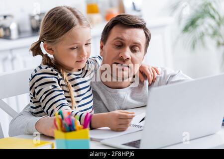 padre che aiuta la figlia a fare i compiti vicino al laptop in primo piano sfocato Foto Stock