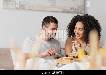 allegra coppia interracial sorridente e che ha fatto colazione in camera da letto Foto Stock