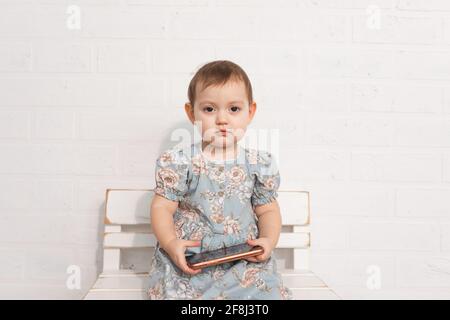 il bambino della piccola ragazza triste si siede sulla panca di legno in abito e tiene lo smartphone nelle sue mani.concetto di sostituzione bambino con genitori con gadget Foto Stock