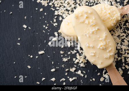 Dessert estivi e spuntini freddi con due gelaterie su un bastone o lecci, circondati da scaglie di cioccolato bianco su una pietra nera scura b Foto Stock