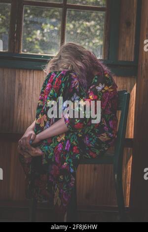 adorabile ragazza con lunghi capelli biondi seduta sulla sedia dietro la finestra e rilassante Foto Stock