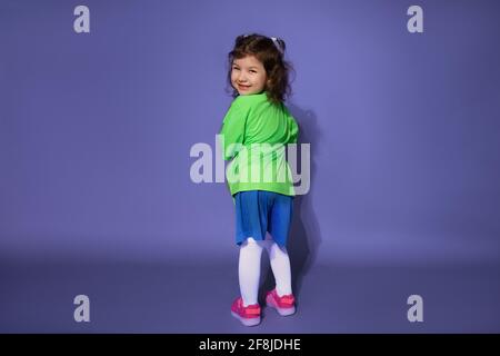 Divertente bambina in uniforme da calcio si alza con la schiena su uno sfondo viola e guarda nella cornice Foto Stock