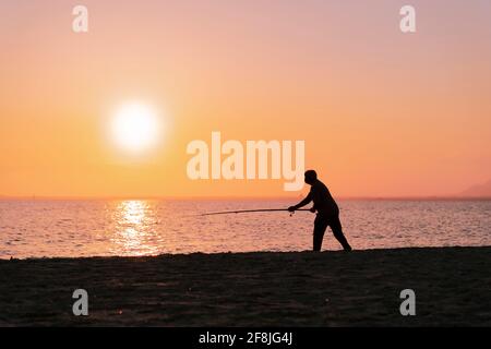 Silhouette di uomo in pesca onde sulla spiaggia al tramonto Foto Stock