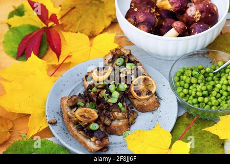 Funghi arrostiti su toast, piselli verdi e funghi crudi (boletus) su sfondo di foglie autunnali (colore verde, giallo e marrone). Foto Stock