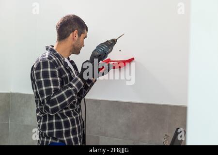 Un falegname che fora la parete per installare i ripiani. Tiene un trapano in una mano, mentre con l'altra tiene una piccola spatola per raccogliere la d Foto Stock