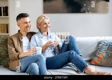 Amante felice adulto sposato caucasico coppia in abiti eleganti casual, felice marito e moglie seduta sul comodo divano nel soggiorno, tenendo tazze di tè, chiacchierando, trascorrere il tempo a casa Foto Stock