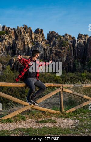 Uomo caucasico spagnolo seduto su recinzione di legno con armata distesa, ammirando la vista della montagna Foto Stock