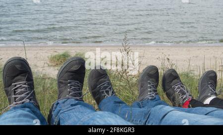 Persone sedute con gambe allungate su una collina, tre coppie di piedi sparsi in sneakers grigio su uno sfondo di erba verde che cresce sulla collina e bl Foto Stock