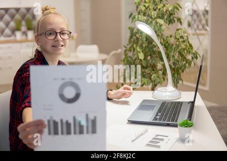 Bionda ragazza con gli occhiali lavoro su un computer portatile e mostra grafici. Concetto di casa-scuola Foto Stock