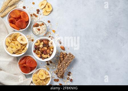 Snack sani, ricchi di vitamine e dolci. Trucioli di banana, albicocche secche, vari tipi di noci, uvetta e barretta di muesli su fondo grigio. Vista dall'alto con Foto Stock