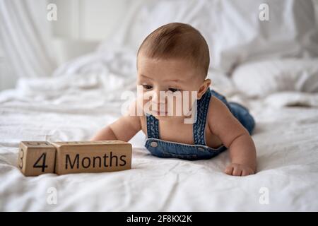 Adorabile bambino di quattro mesi in denim dungarees mentendo su letto bianco Foto Stock