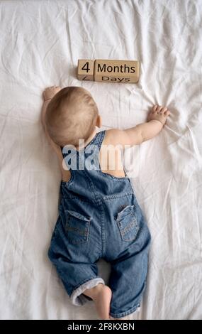 Adorabile bambino di quattro mesi in denim dungarees mentendo su letto bianco Foto Stock