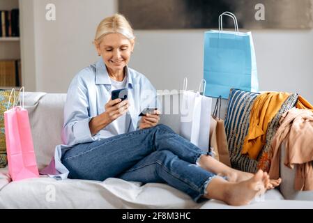 Gioiosa bella caucasica matura bionda donna elegante, felice con i nuovi vestiti acquistati, seduta su un divano a casa, utilizzando smartphone e carta di credito per i pagamenti online, molti sacchetti di carta vicino, sorridente Foto Stock