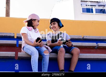 Bambini caucasici seduti sui bleachers con i loro dispositivi mobili e le cuffie, guardandosi e sorridendo molto felicemente. Foto Stock