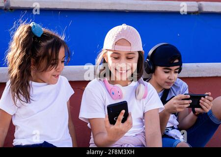 Bambini caucasici seduti sui bleachers con i loro dispositivi mobili e le cuffie, condividendo momenti di risate e conversazioni. Foto Stock