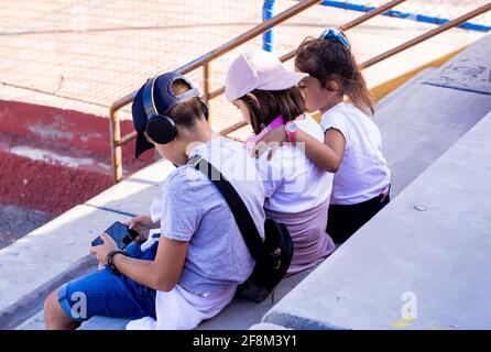 Bambini caucasici seduti sui bleachers con i loro dispositivi mobili e le cuffie, condividendo momenti di risate e conversazioni. Foto Stock