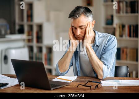 Uomo, manager o freelance, spossato dai capelli grigi caucasici, seduto alla scrivania, tenendo la testa con le mani, stanco di lavorare in un computer portatile, ha mal di testa, emicrania, ha bisogno di riposo Foto Stock