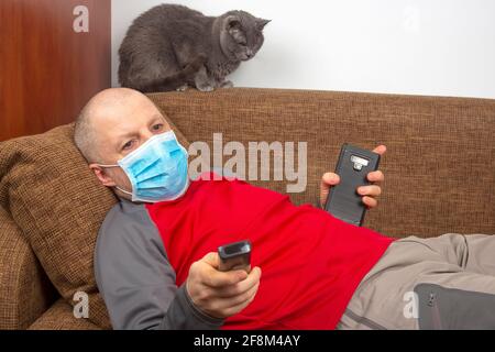 uomo in quarantena a casa con una maschera medica su il suo volto si trova sul divano e guarda le notizie In TV accanto a un gatto grigio Foto Stock