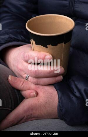 La mano della donna tiene la tazza di carta con il caffè, l'altra mano si appoggia sulle gambe. Mettere a fuoco la mano superiore e il bordo della tazza marrone. Foto verticale Foto Stock