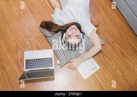 Vista dall'alto di una giovane bella donna asiatica con cuffie e ascolto di musica dal computer portatile mentre si trova sul pavimento a casa. Foto Stock