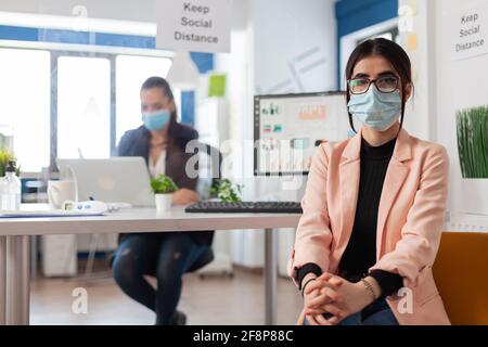 Ritratto del lavoratore dell'ufficio dell'azienda che indossa la maschera facciale come precauzione di sicurezza durante la pandemia globale con il coronavirus che osserva la macchina fotografica che mantiene la distanza sociale. Responsabile che lavora su un computer portatile con una pellicola in plastica sulla scrivania. Foto Stock